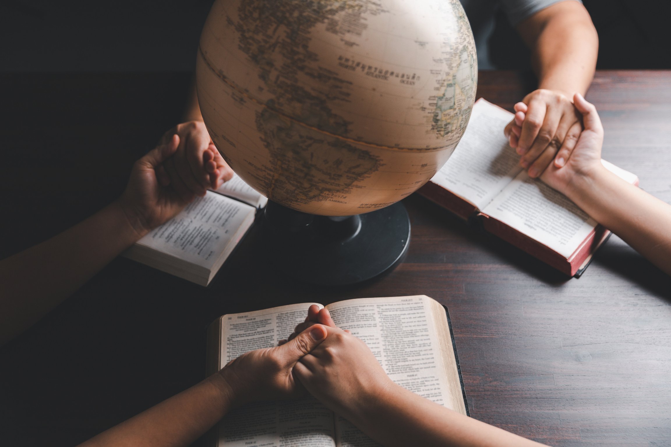 Christian group praying for globe and people around the world on wooden table with bible. Christian small group holding hands and praying together around a wooden table with bible page in homeroom.